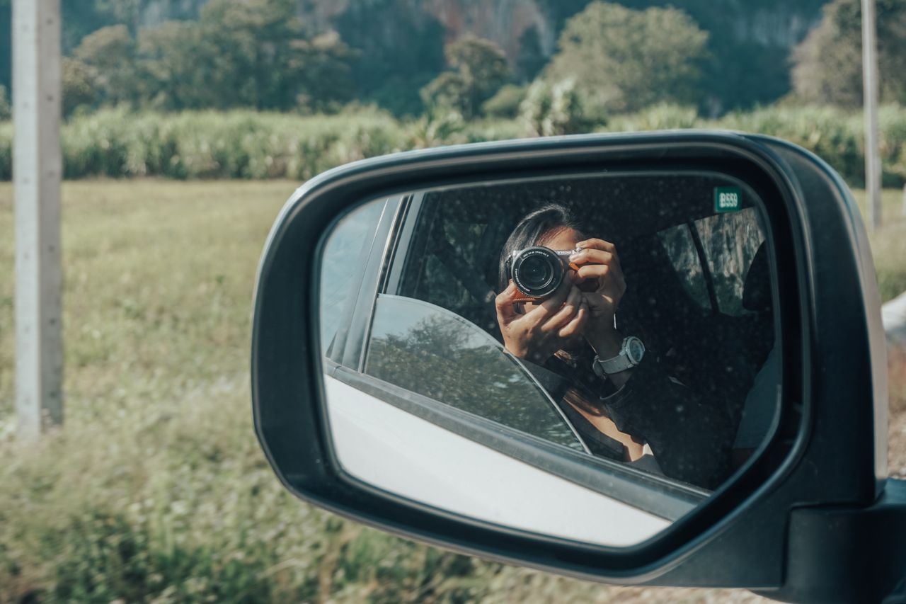 A woman captures the scenic countryside through a side mirror reflection, showcasing travel and photography.