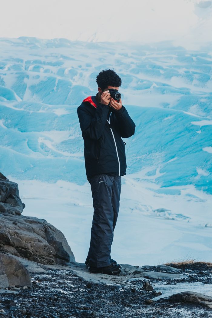 Faceless male camper standing on stony ground near snowy surface and taking photos of scenery in winter time in daytime